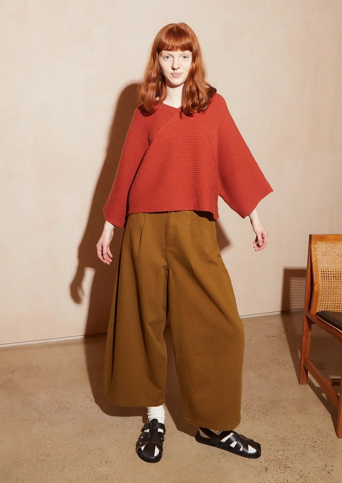 A model with red hair stands in a studio wearing a rust-orange ribbed knit poncho-style top and wide-leg brown trousers, paired with white socks and black sandals. The background is a warm beige wall and floor with a wooden chair partially visible on the right.