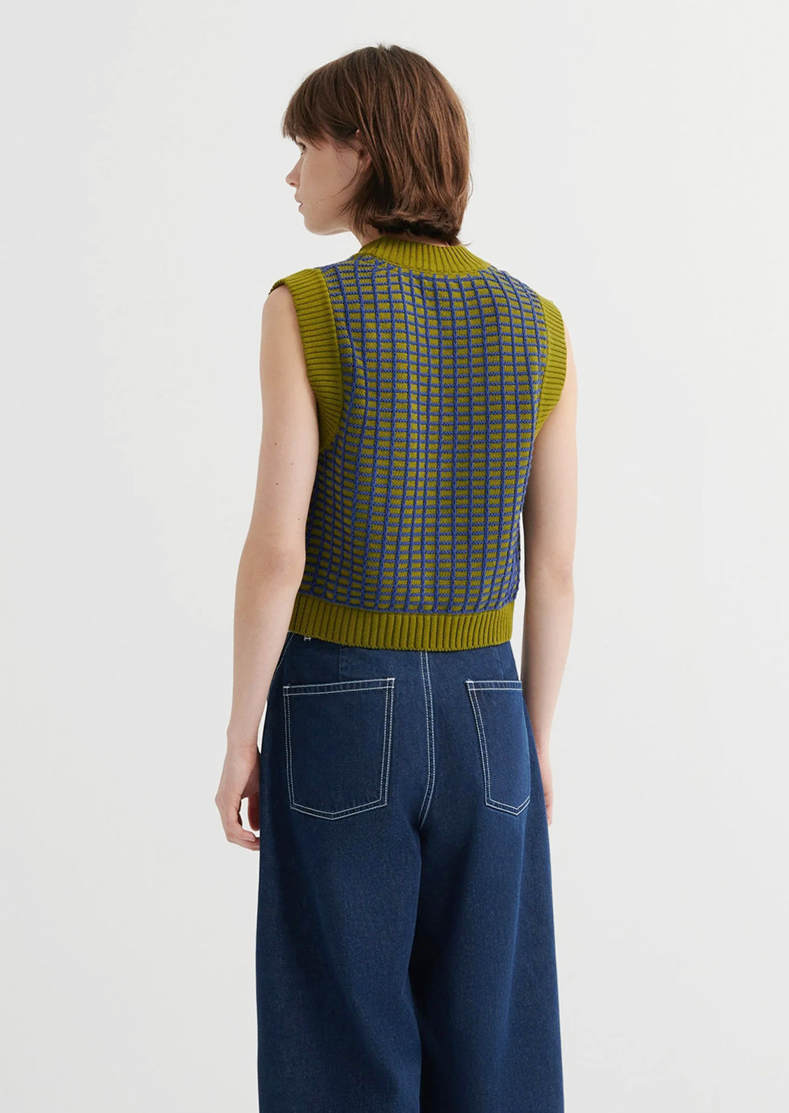 Person wearing a green checkered sleeveless top and blue jeans on a white background