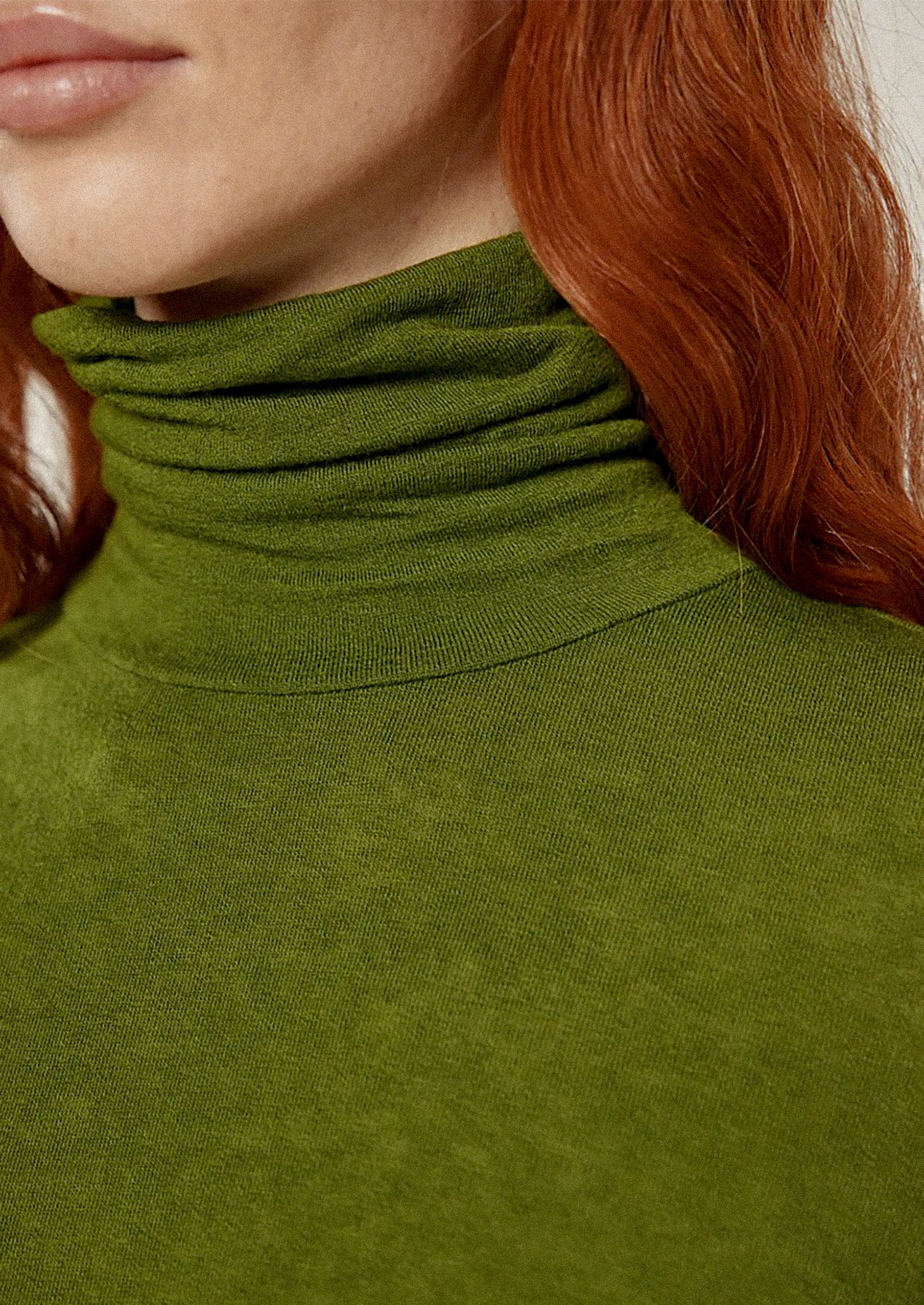 Person wearing a green turtleneck sweater and brown pants against a white background