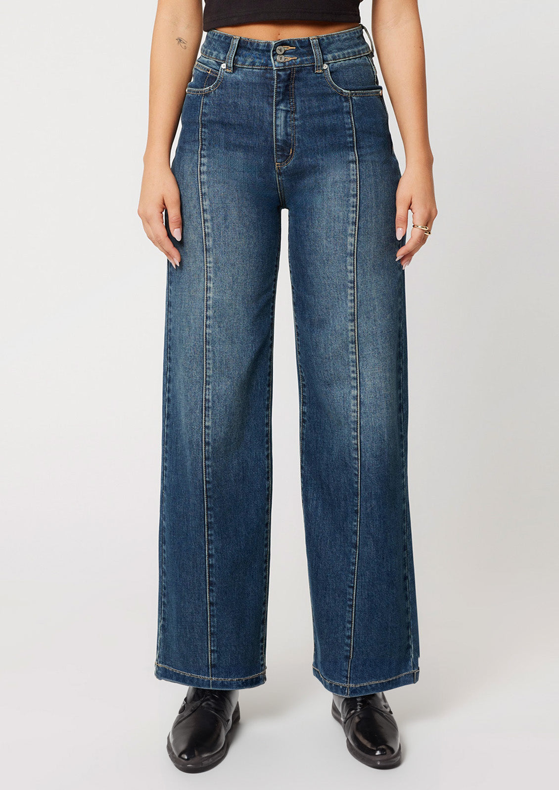 A woman wearing a pair of wide leg, high rise jeans in medium dark blue wash with seam down center leg.