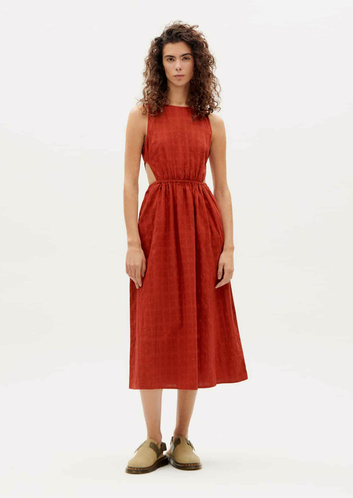 A woman wearing a rust-hued sleeveless dress with tonal square texture and cutout detail at waist.