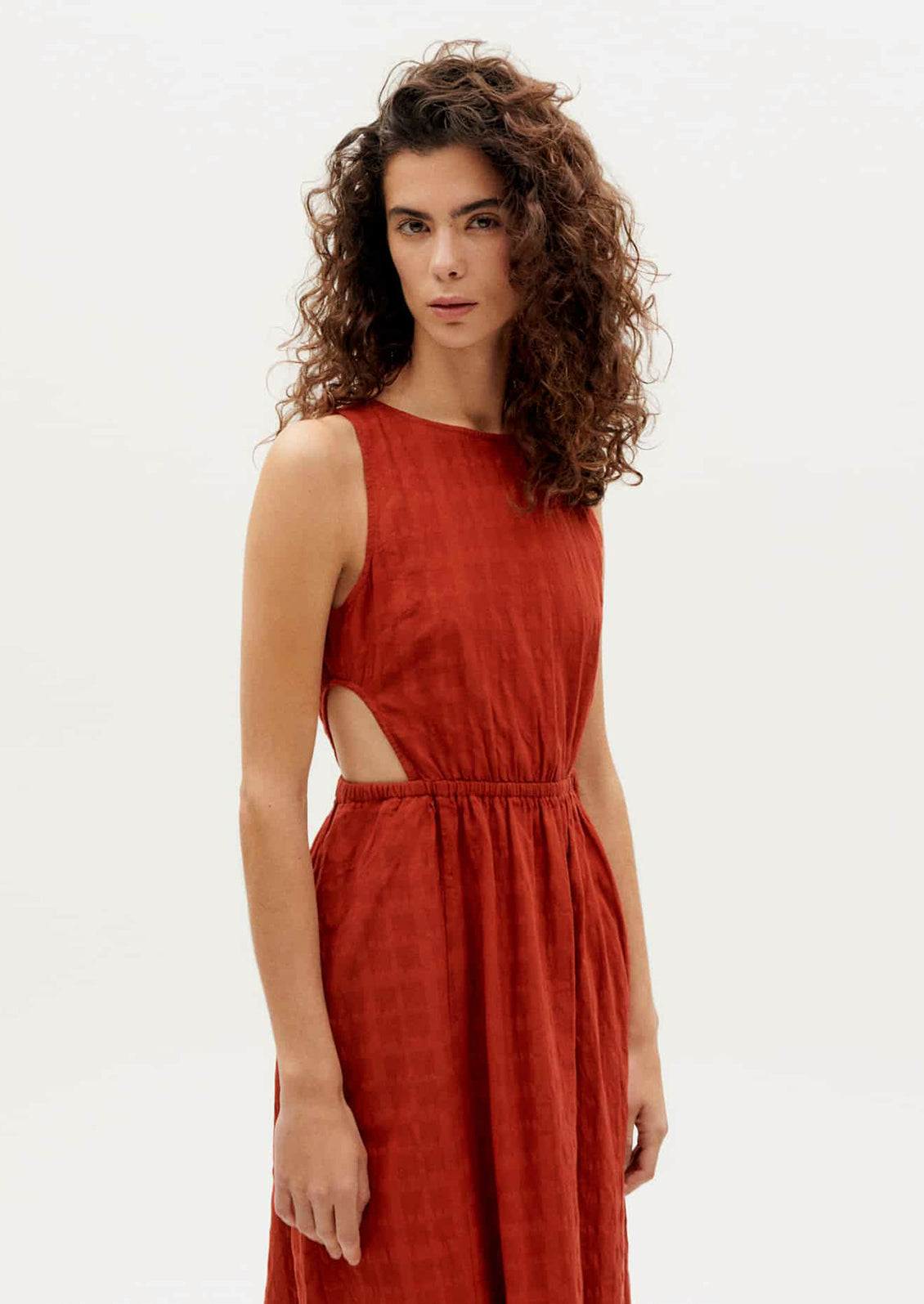 A woman wearing a rust-hued sleeveless dress with tonal square texture and cutout detail at waist.