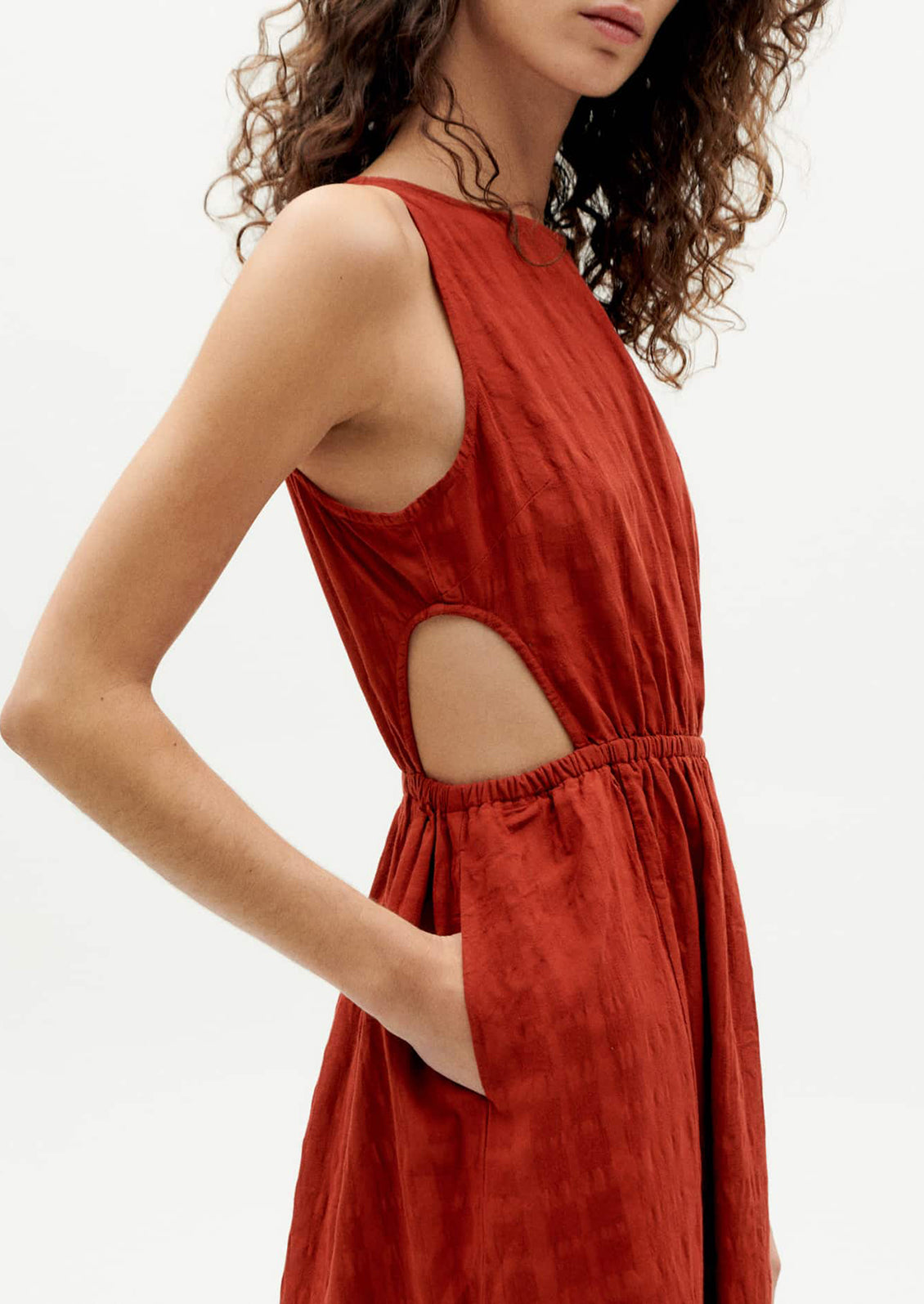 A woman wearing a rust-hued sleeveless dress with tonal square texture and cutout detail at waist.