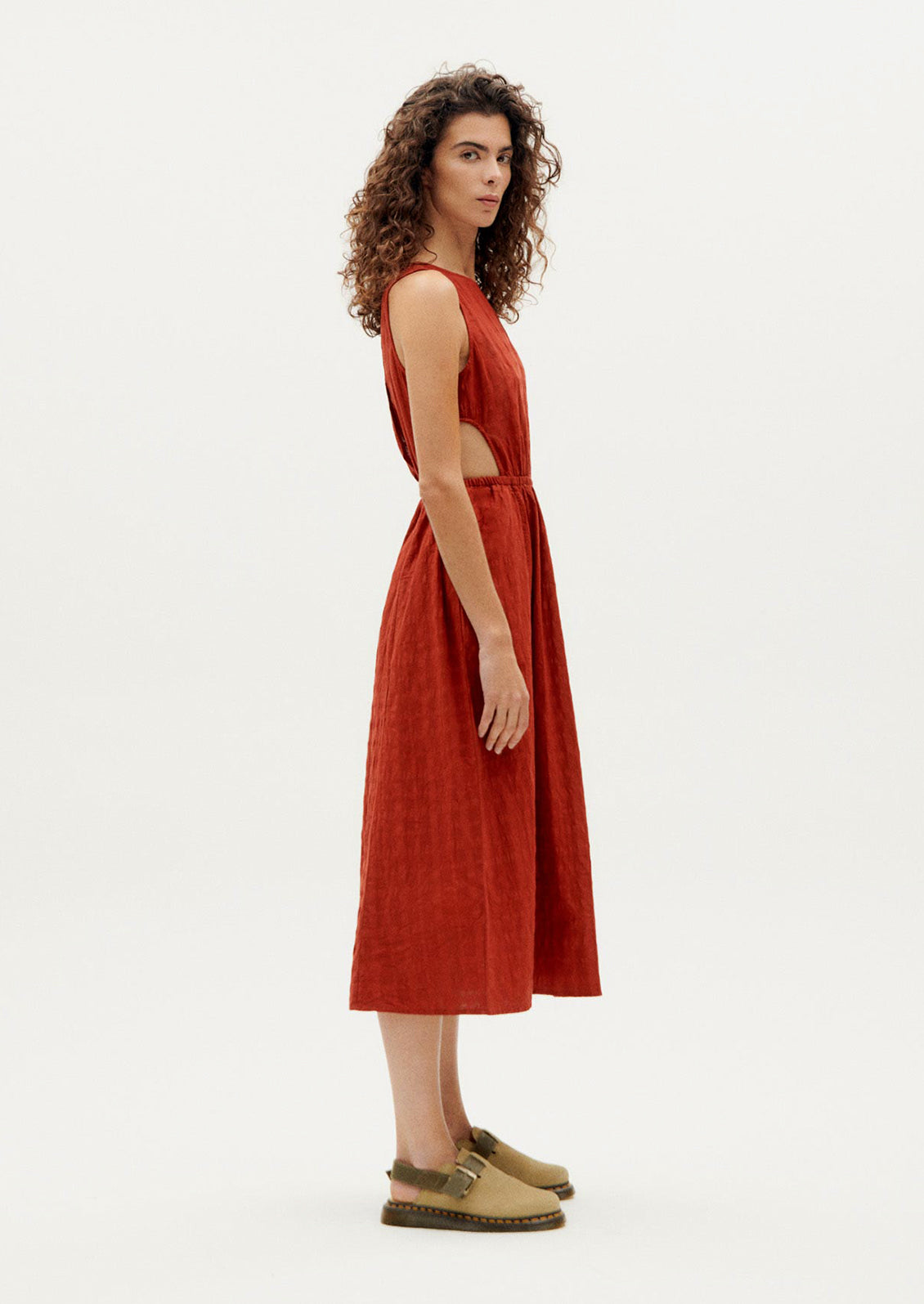 A woman wearing a rust-hued sleeveless dress with tonal square texture and cutout detail at waist.