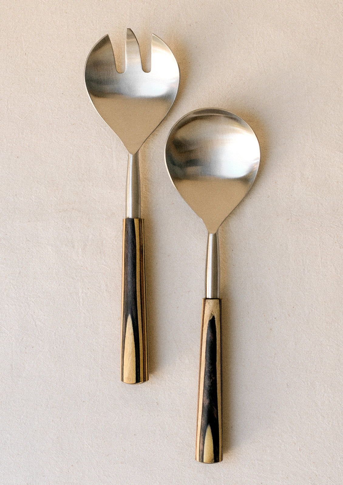A pair of stainless steel, modern salad servers with two-tone wooden handle.