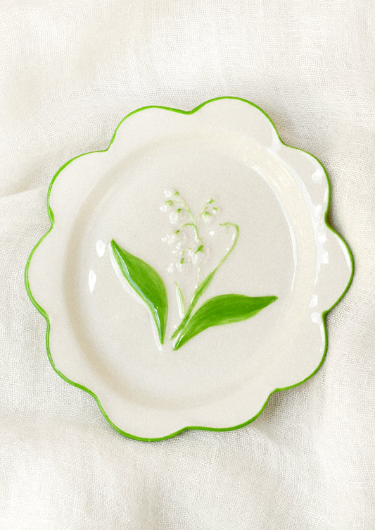 A ceramic plate with scalloped edges and a lily of the valley embossed in the center.