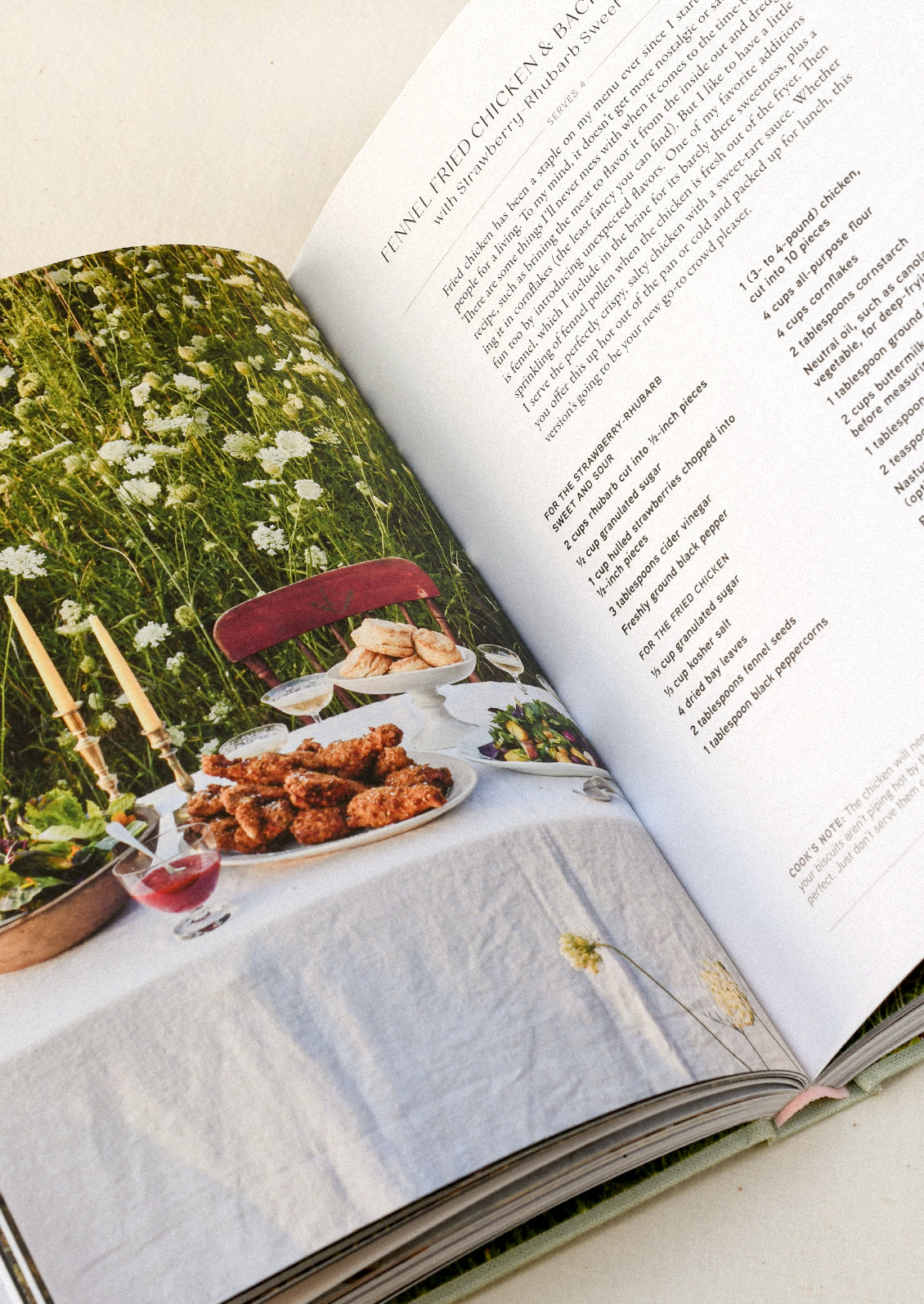 A cookbook open on a table showing a recipe for fennel fried chicken on one page and an image of a dining table set up outside on the other.