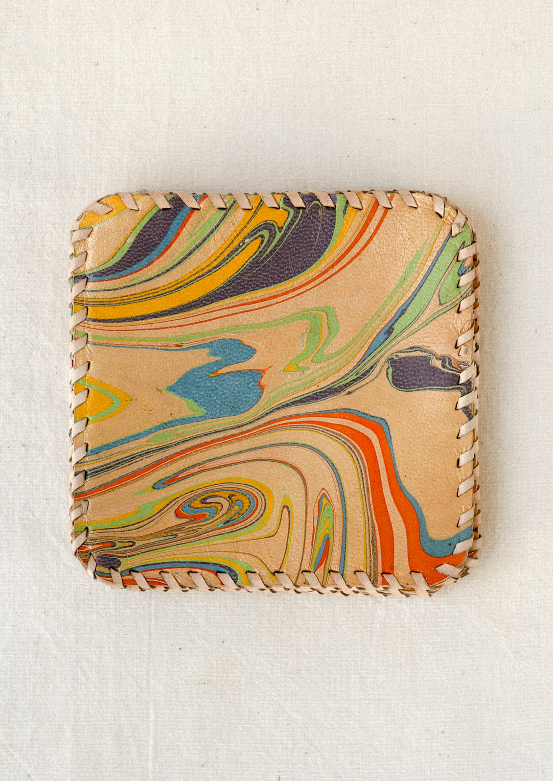 Square leather coaster with rounded corners, featuring a colorful swirling marbled pattern in mustard yellow, green, blue, orange, and purple. The edges are finished with contrasting cream-colored whipstitching.