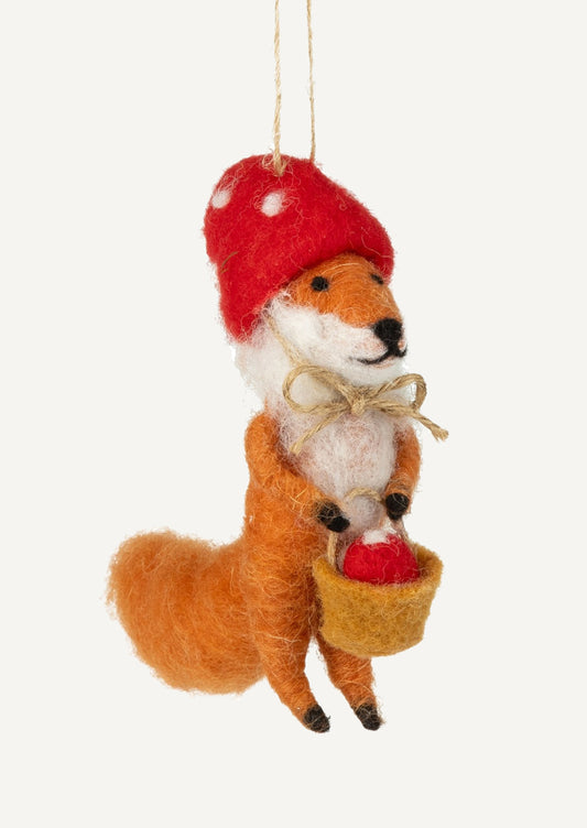 A felted wool ornament of fox wearing mushroom cap tied in a bow and carrying basket of mushrooms.