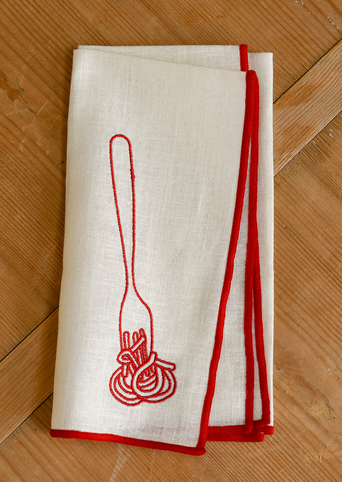 An ivory cloth dinner napkin with red edging, and red embroidery of spaghetti on fork.
