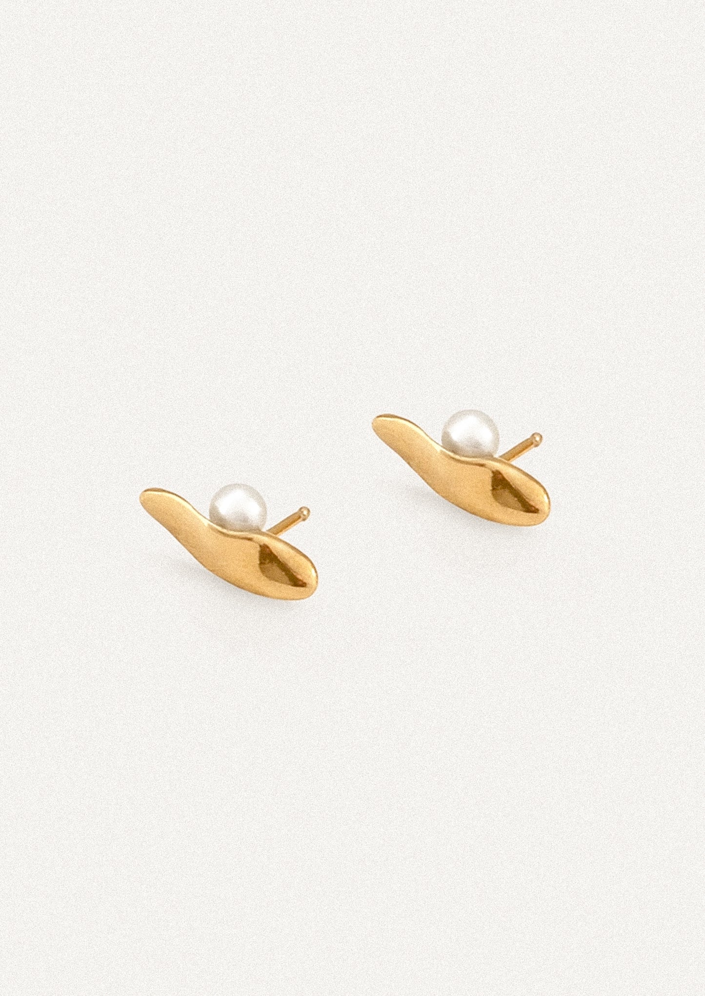 A pair of gold-toned stud earrings shaped like stylized leaves or teardrops, each set with a small white pearl, shown on a clean off-white background.