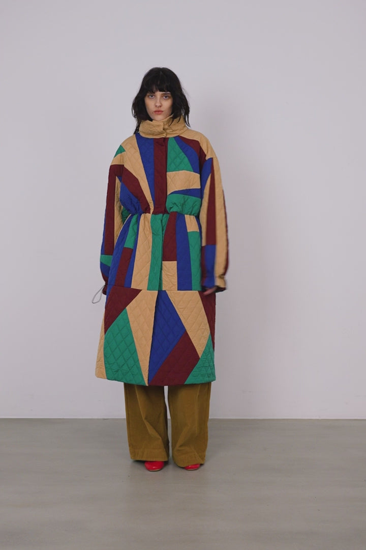 A woman wearing a geometric patchwork quilted coat in wine, teal, beige and blue.