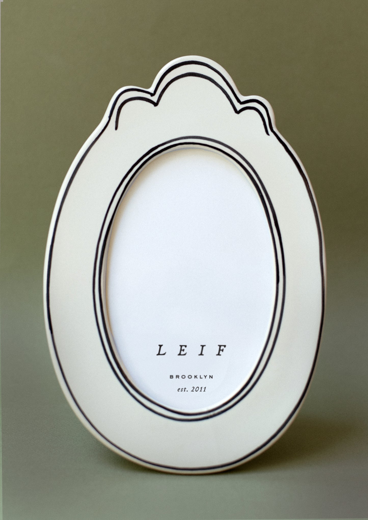 A white ceramic picture frame with ornate oval shape and thin handpainted border detail.