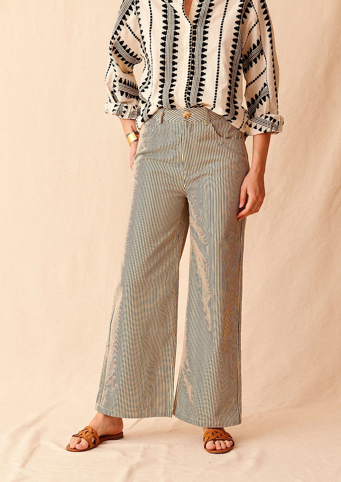 A woman standing wearing cream and black pinstriped wide leg jeans with brown sandals and cream and black embroidered top