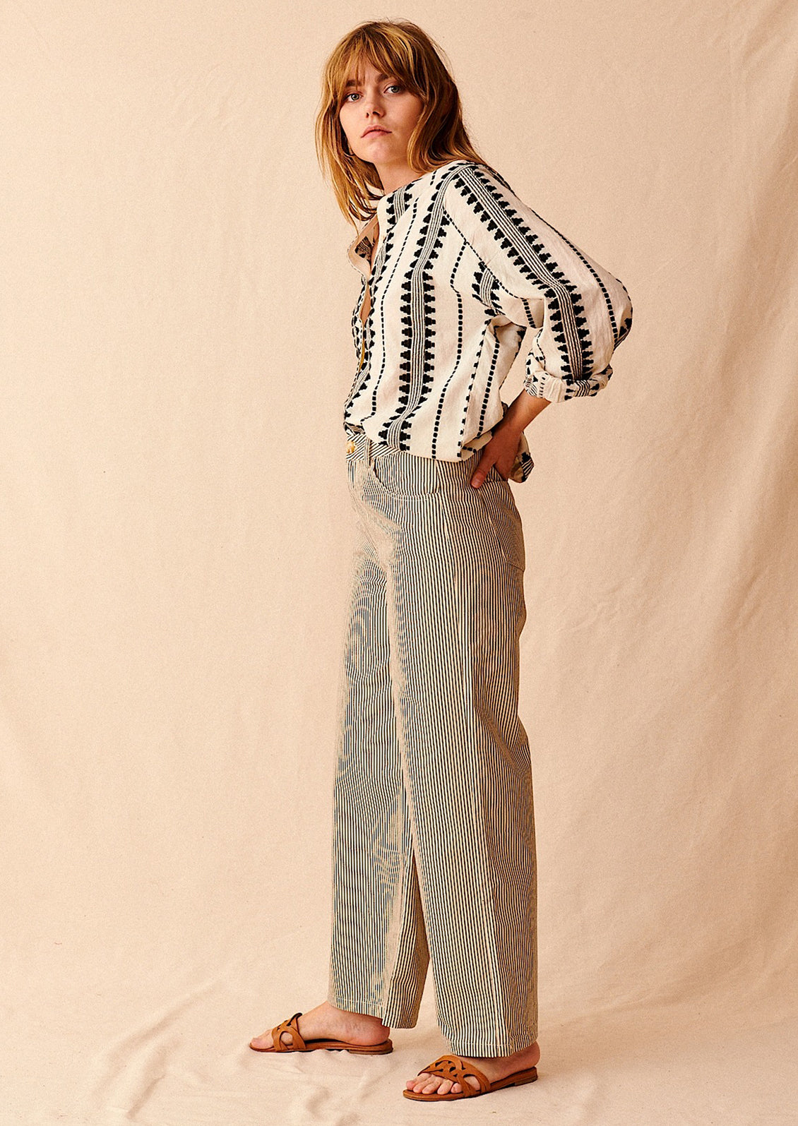 A woman standing wearing cream and black pinstriped wide leg jeans with brown sandals and cream and black embroidered top, seen from side
