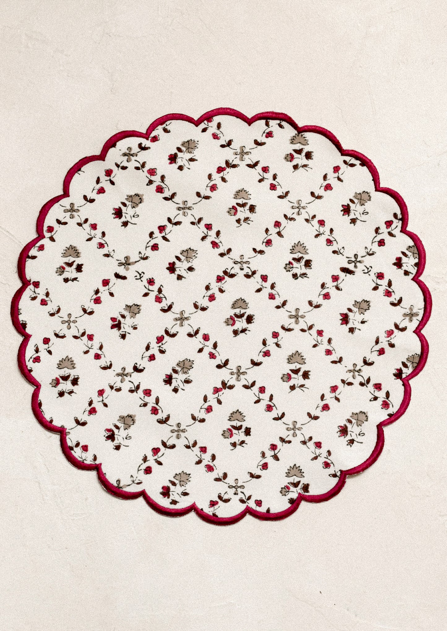 A round scalloped placemat in white with tan and burgundy floral print and embroidered burgundy trim.