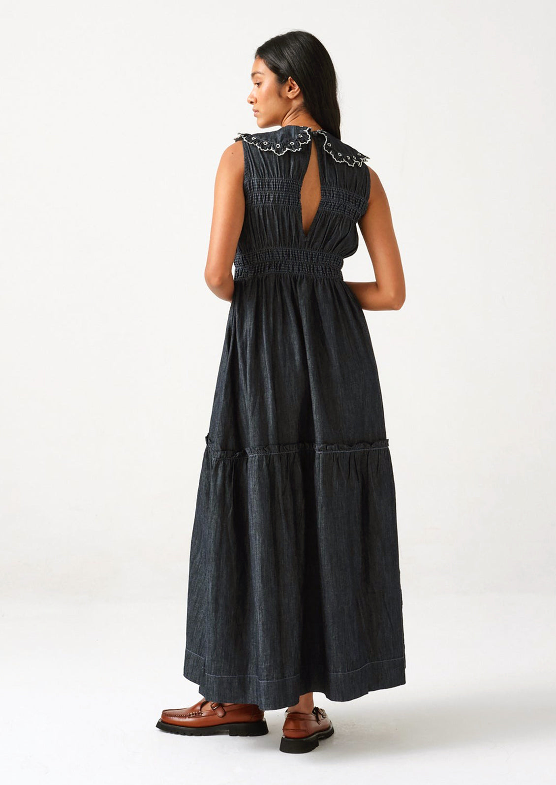 A woman standing wearing a long denim dress with an embroidered collar, viewed from back.