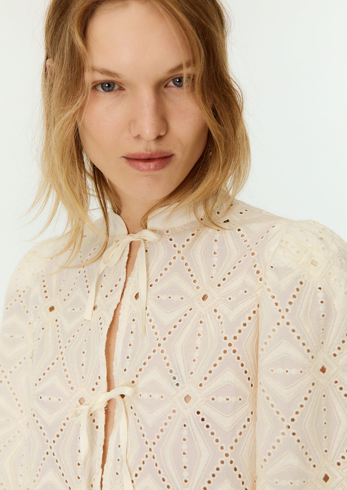 A woman wearing a cream lace blouse with mandarin collar and triple bow tie closure at front.