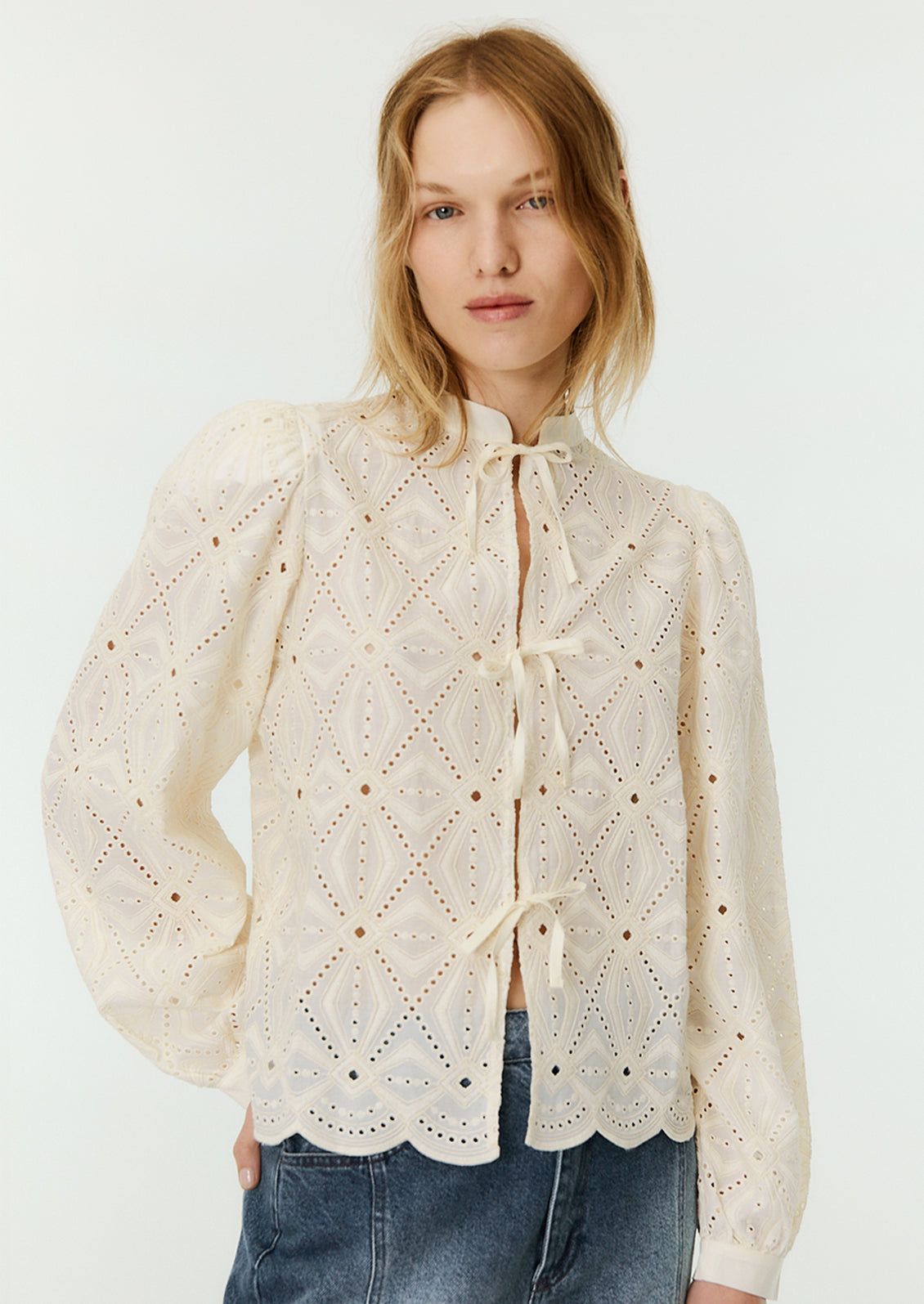 A woman wearing a cream lace blouse with mandarin collar and triple bow tie closure at front.