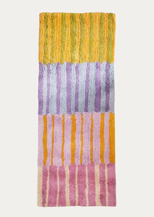 A rectangular plush bath mat with vertical striped blocks in yellow, lavender, pink, and cream, shown flat against a light gray background.