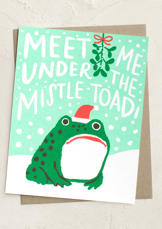 A Christmas greeting card featuring a green toad wearing a red Santa hat, with the text 'MEET ME UNDER THE MISTLE-TOAD!' and a mistletoe sprig on a mint green background, shown with a brown envelope behind it.