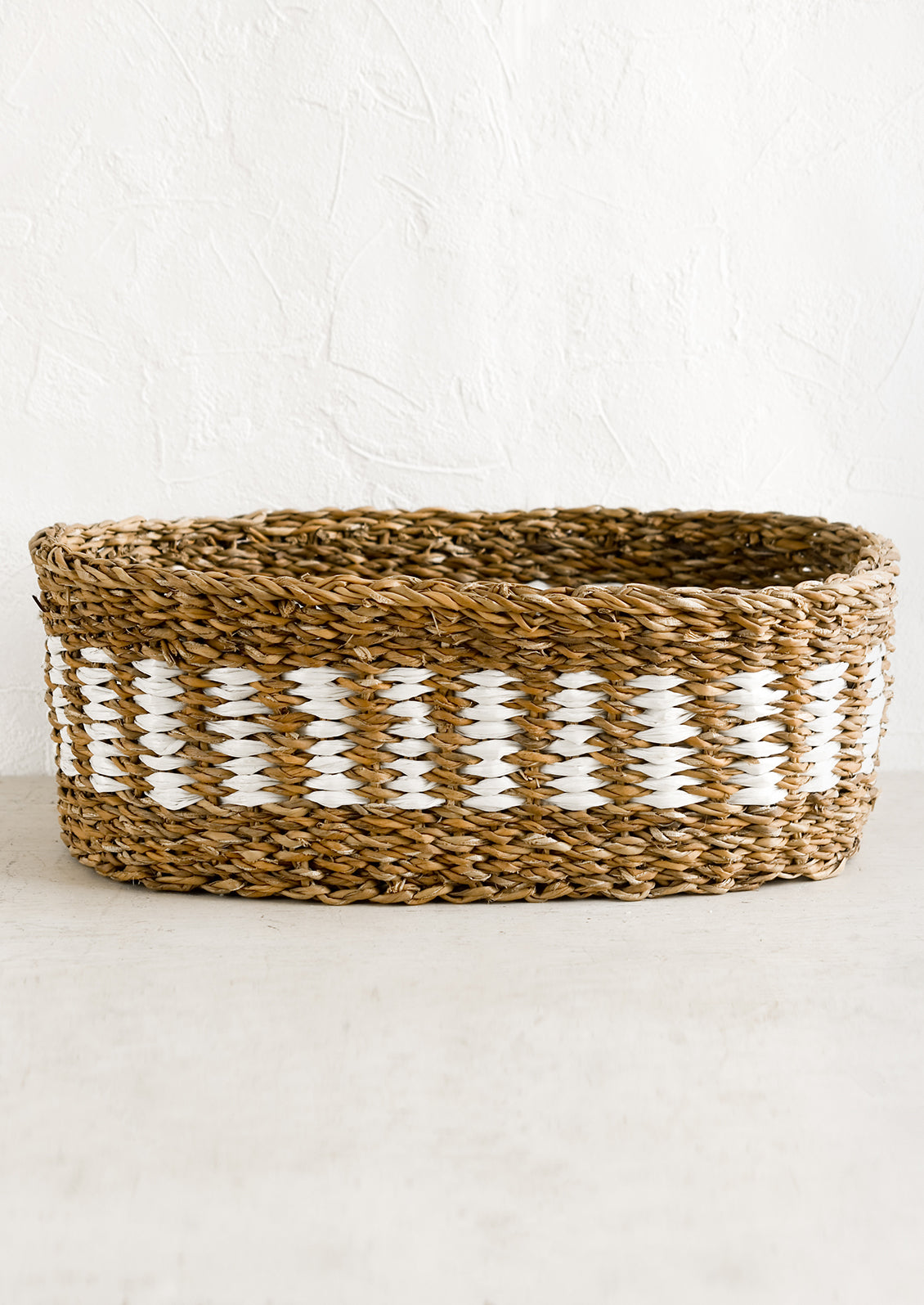 An oval shaped storage basket in seagrass with woven white detail.