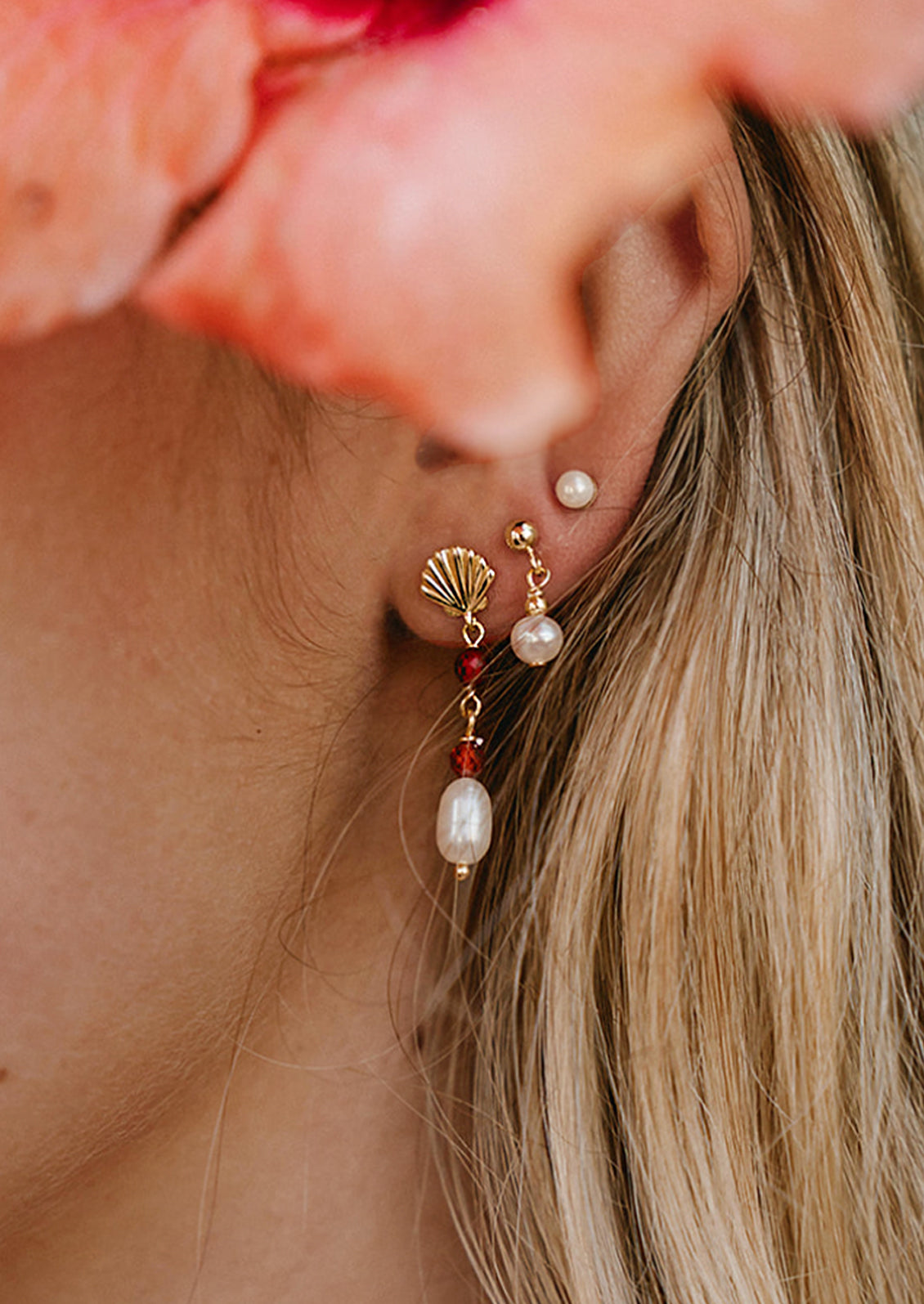 A woman wearing a gold seashell earring with dangling pearl and red gems.
