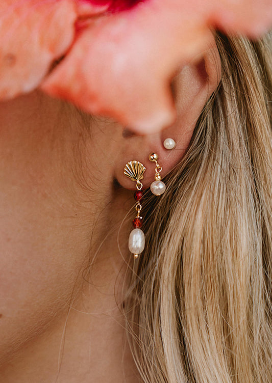 A woman wearing a gold seashell earring with dangling pearl and red gems.