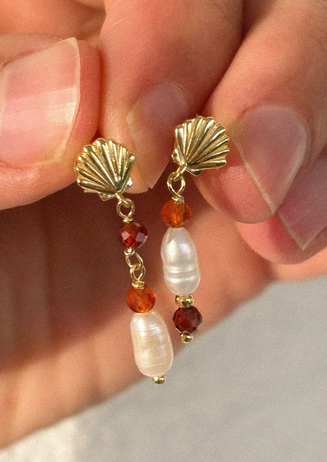 A pair of gold seashell earrings with dangling pearl and red gems.
