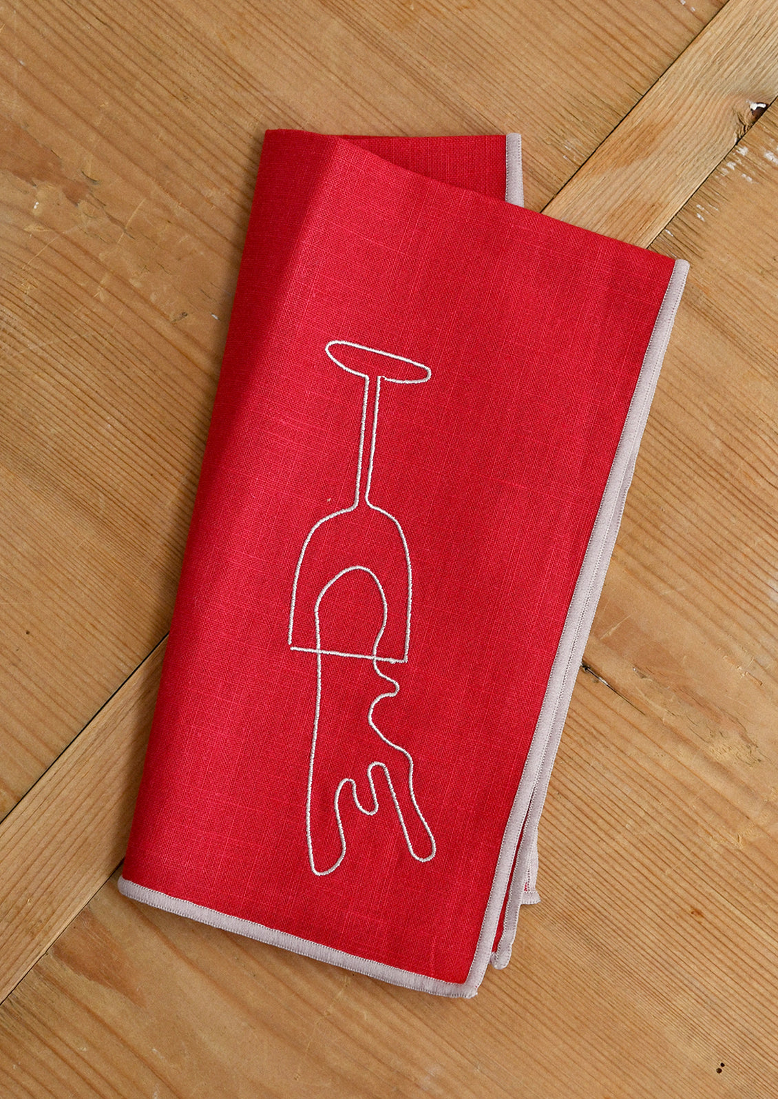 An red cloth dinner napkin with tan edging, and tan embroidery of a wine glass upside down with wine spilling out.