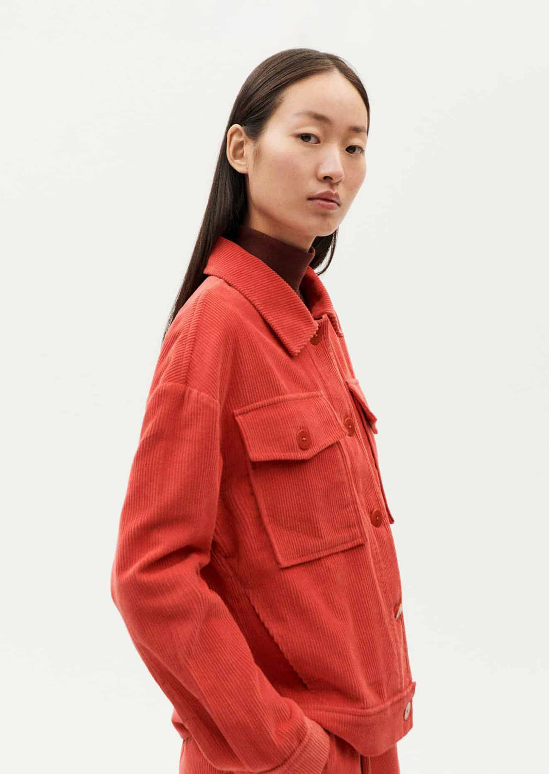 A woman wearing a shirt jacket style corduroy long sleeve jacket in red-orange hue.