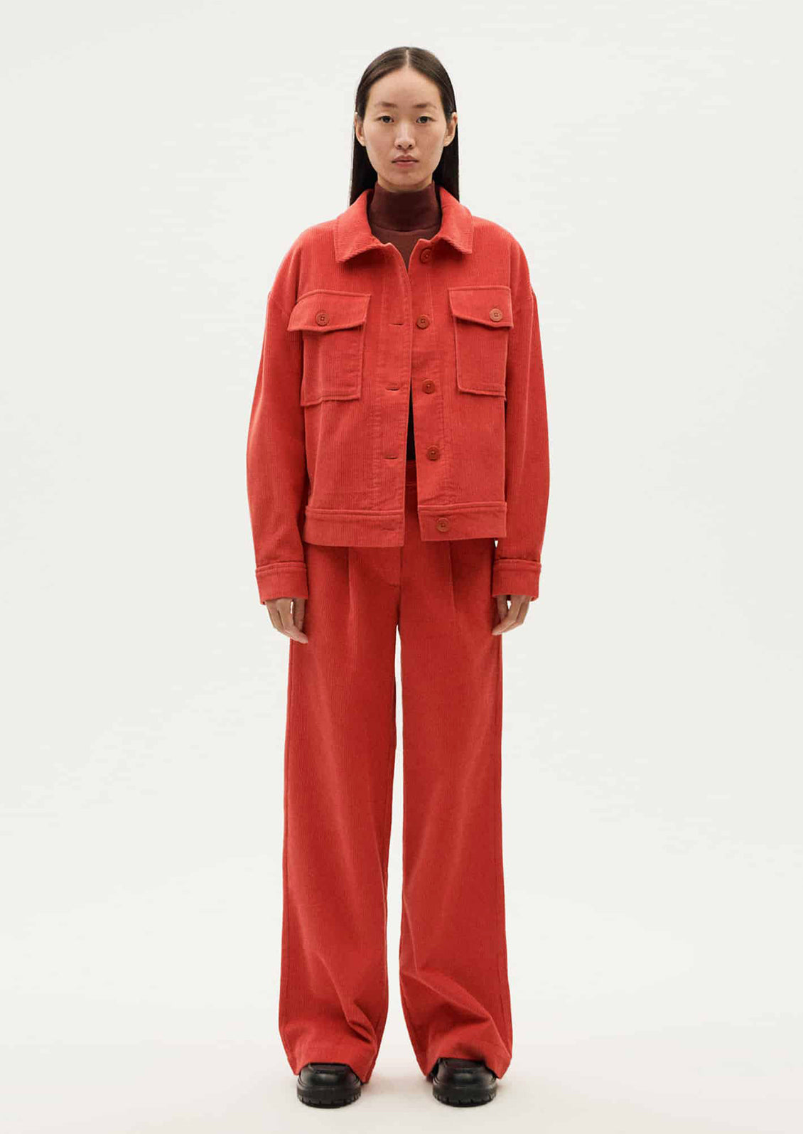 A woman wearing a shirt jacket style corduroy long sleeve jacket in red-orange hue.