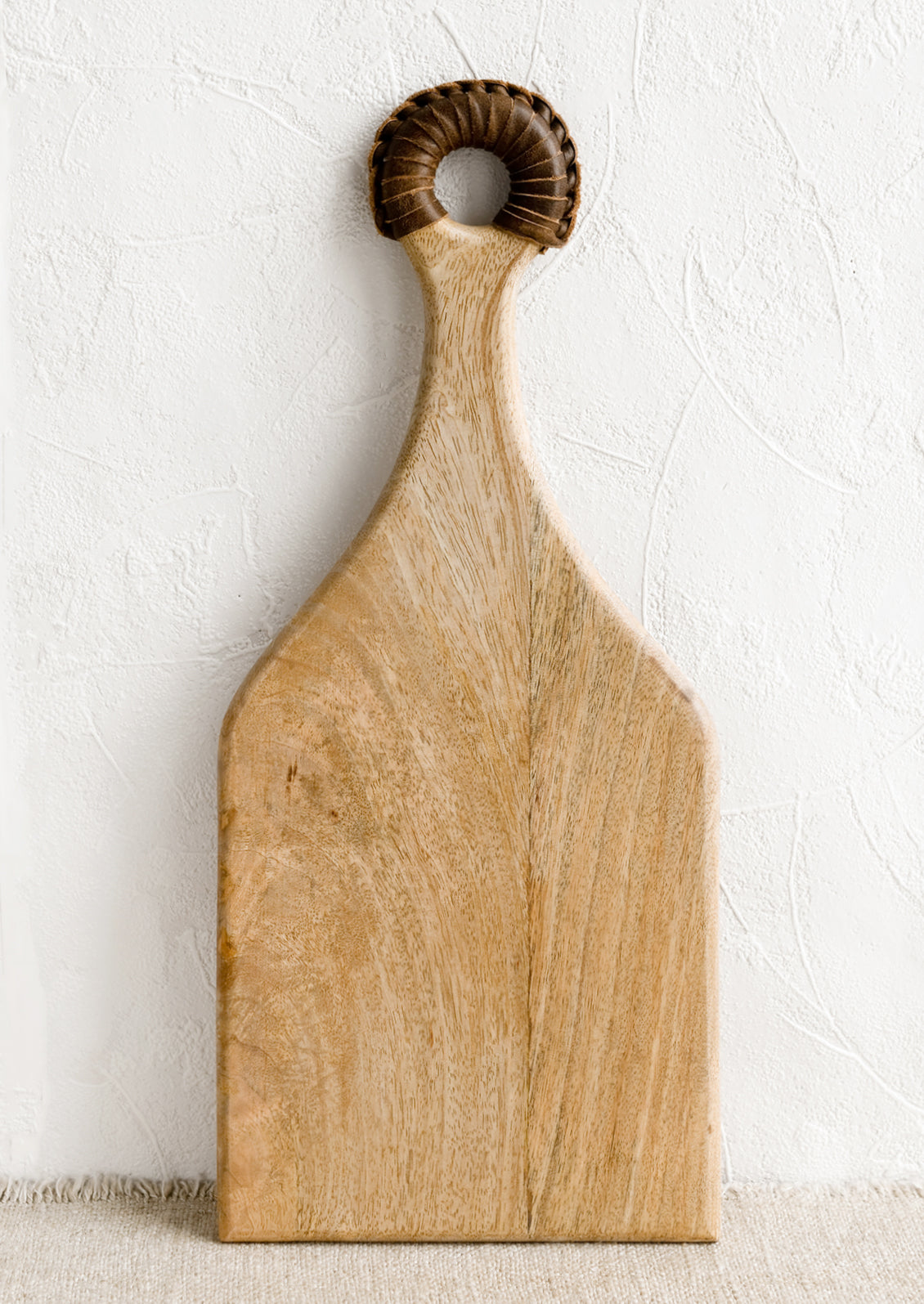 A paddle shaped light wood cutting board with brown leather wrapped handle.
