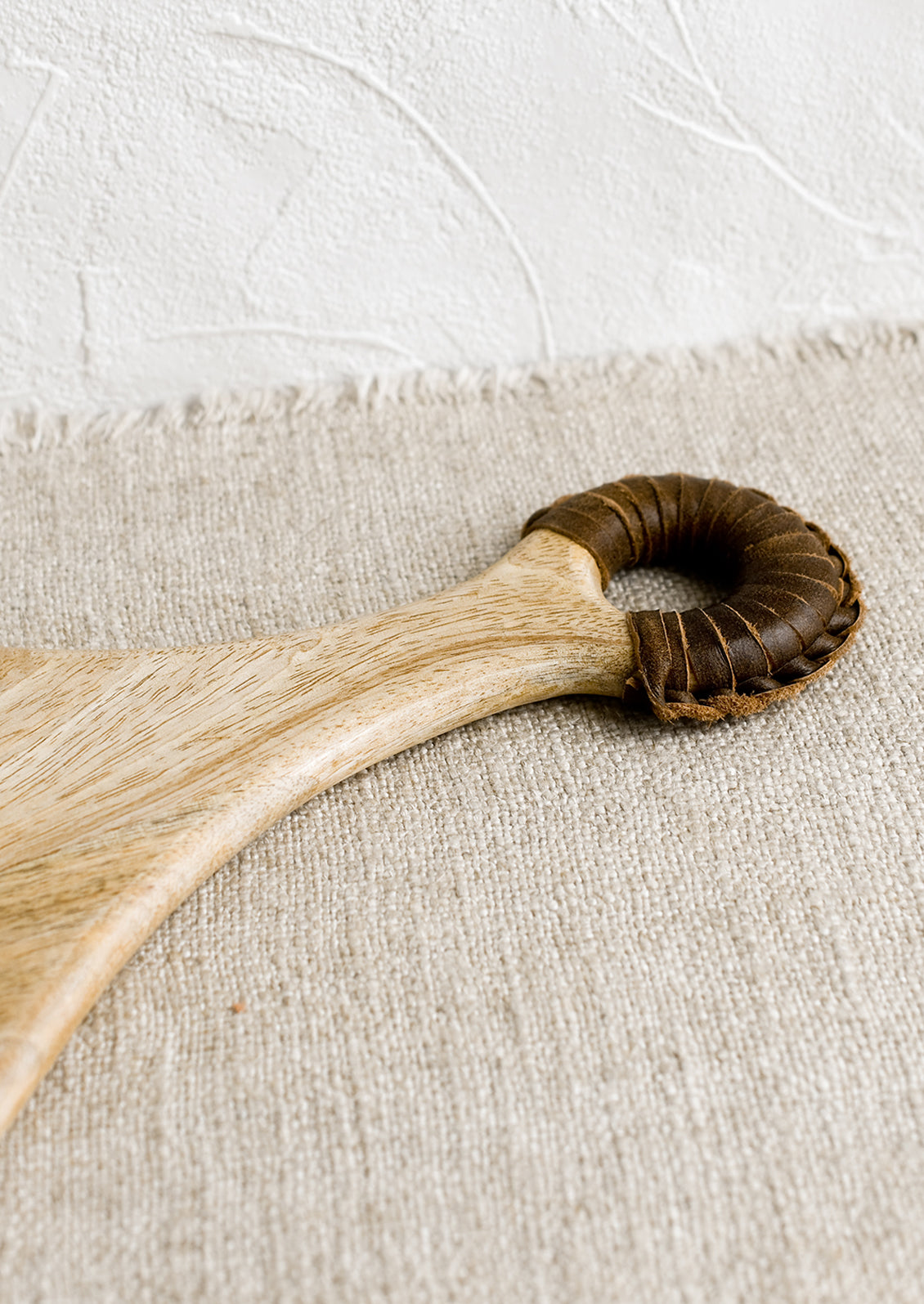 A paddle shaped light wood cutting board with brown leather wrapped handle.