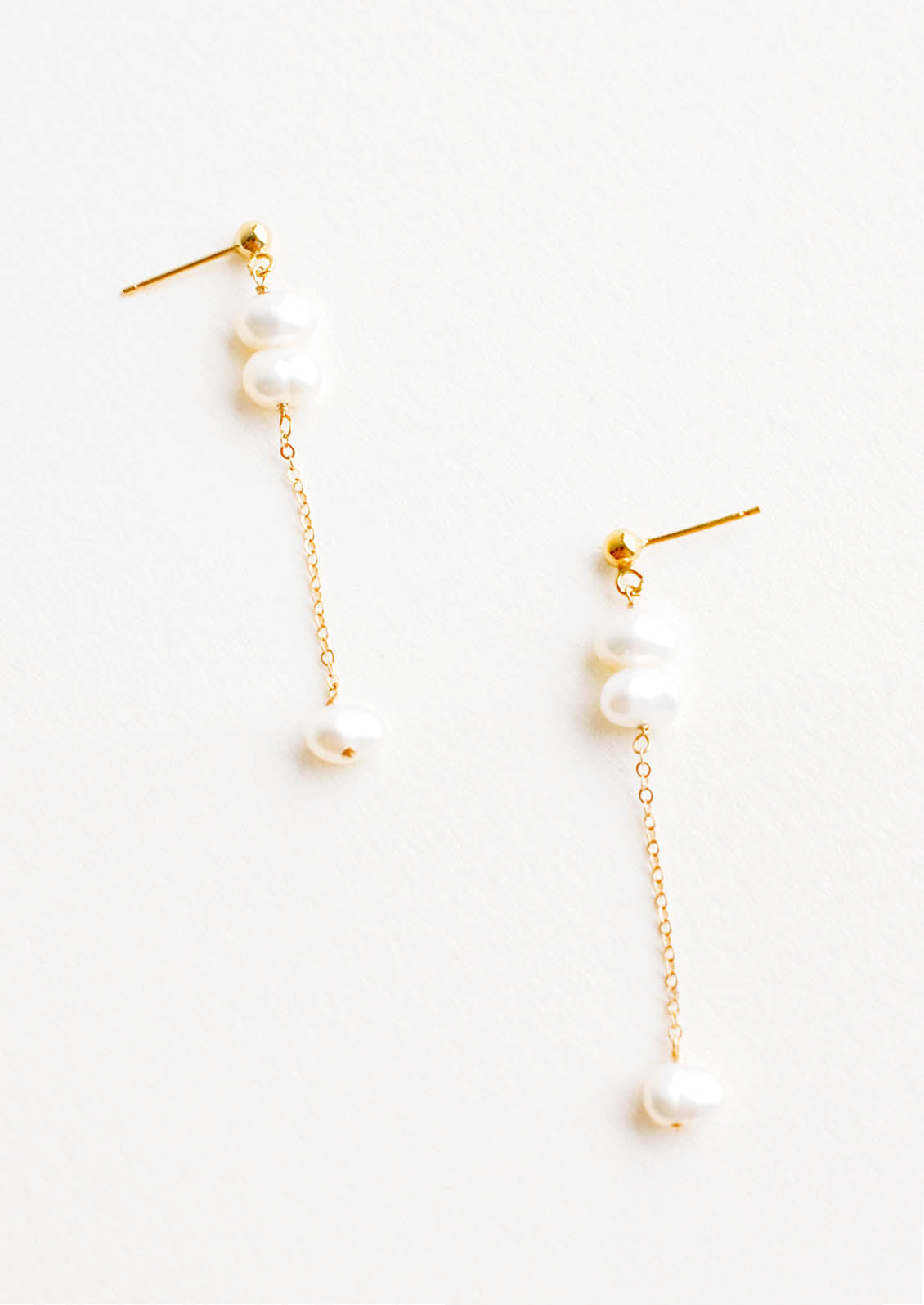 A pair of gold earrings with three pearls dangling from a post, and the third pearl is hanging from a fine gold chain.