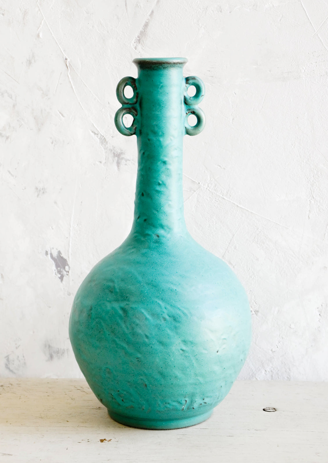 Tall, sculptural ceramic vase in turquoise color with bulbous base and skinny neck. Decorative loop detailing at top sides.