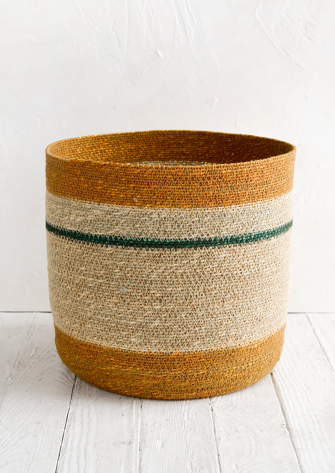 A round open-top storage basket woven from seagrass in ochre, natural and teal stripes.