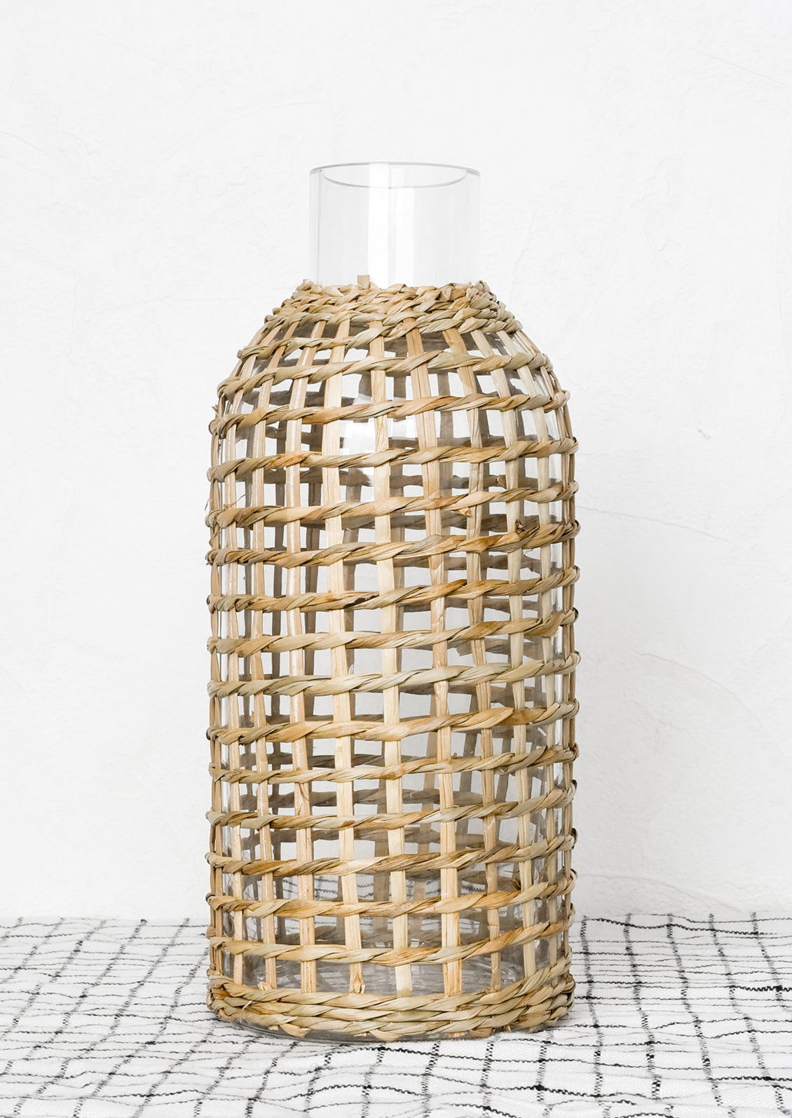 A narrow mouth clear glass bottle wrapped in gridded seagrass.