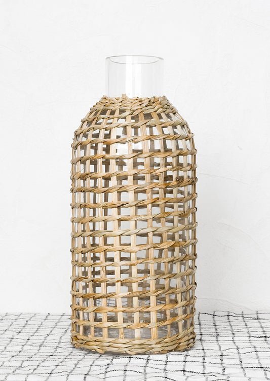 A narrow mouth clear glass bottle wrapped in gridded seagrass.