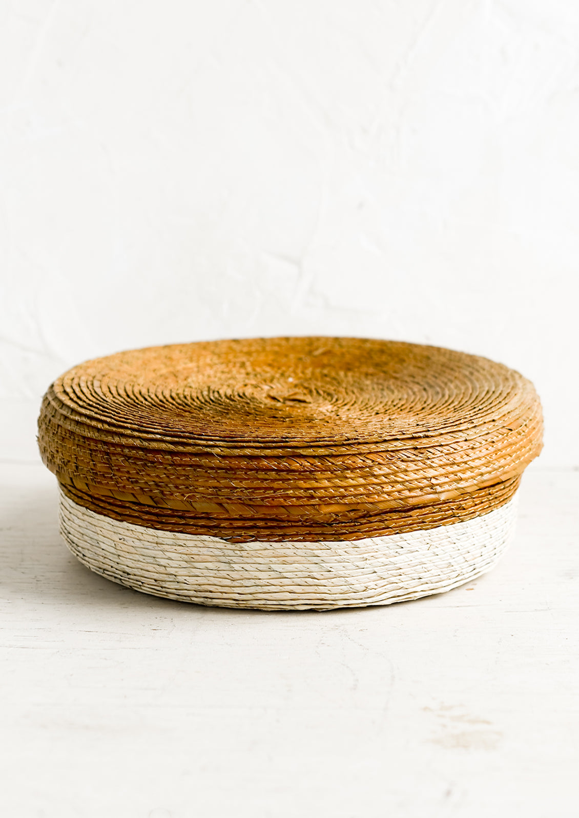 A shallow, round storage basket with lid, woven from dried palm leaf in two-tone design.
