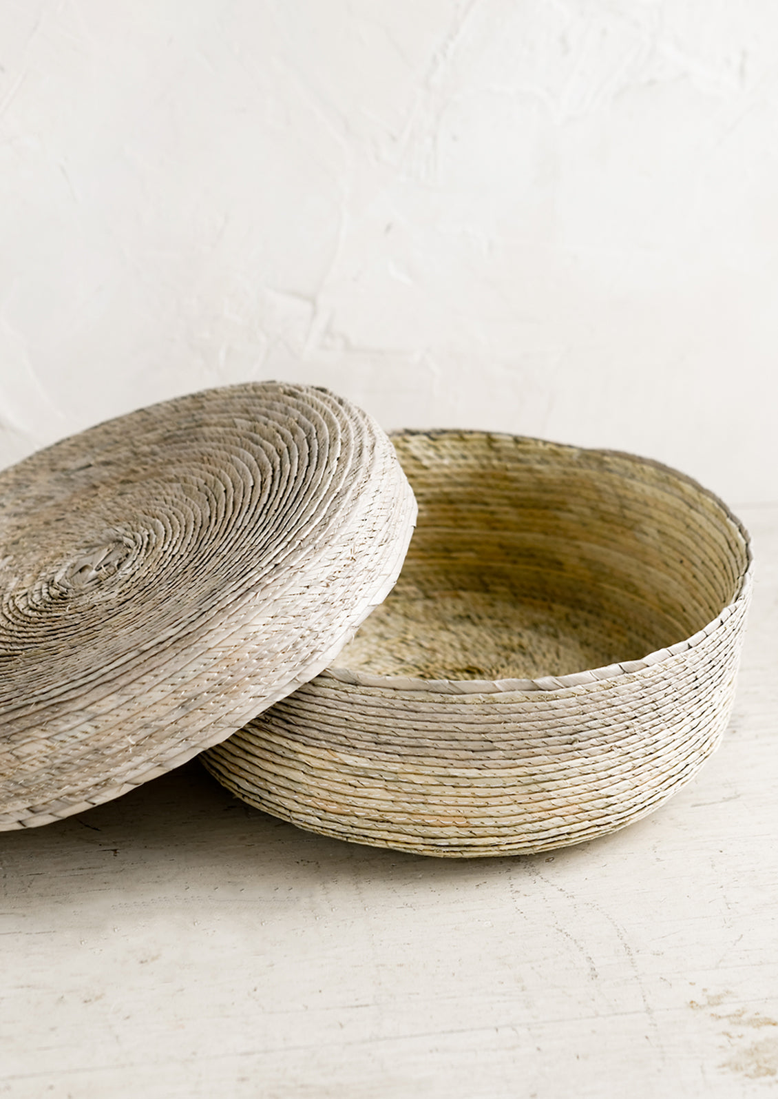 A shallow, round storage basket with lid, woven from dried palm leaf in two-tone design.