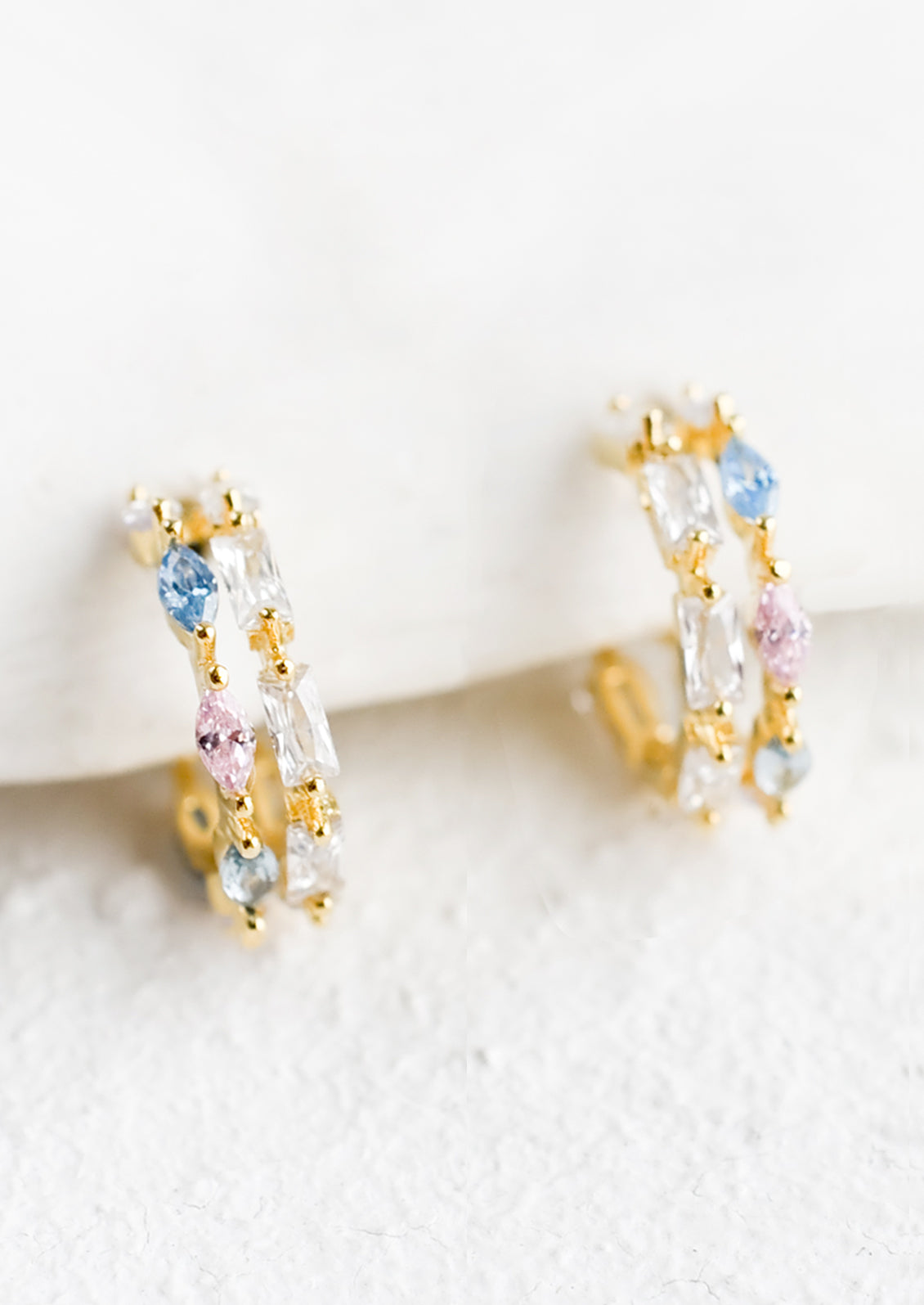 A pair of small gold huggie hoop earrings with clear, pink and blue crystals.