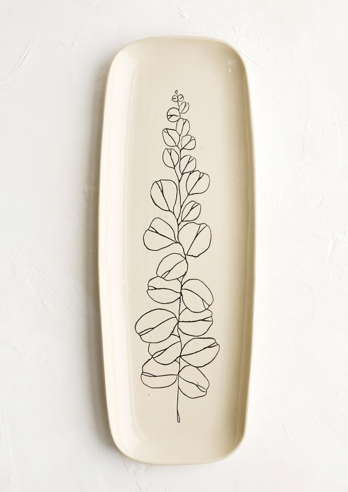 A long and skinny ceramic tray in natural bisque color with an etched black drawing of a eucalyptus branch.