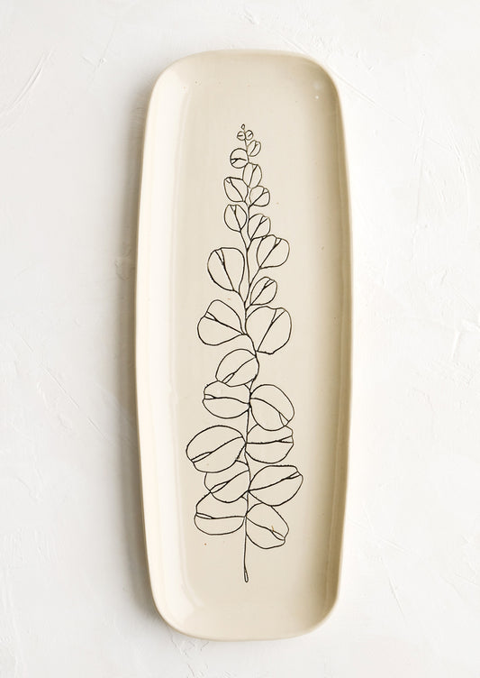 A long and skinny ceramic tray in natural bisque color with an etched black drawing of a eucalyptus branch.