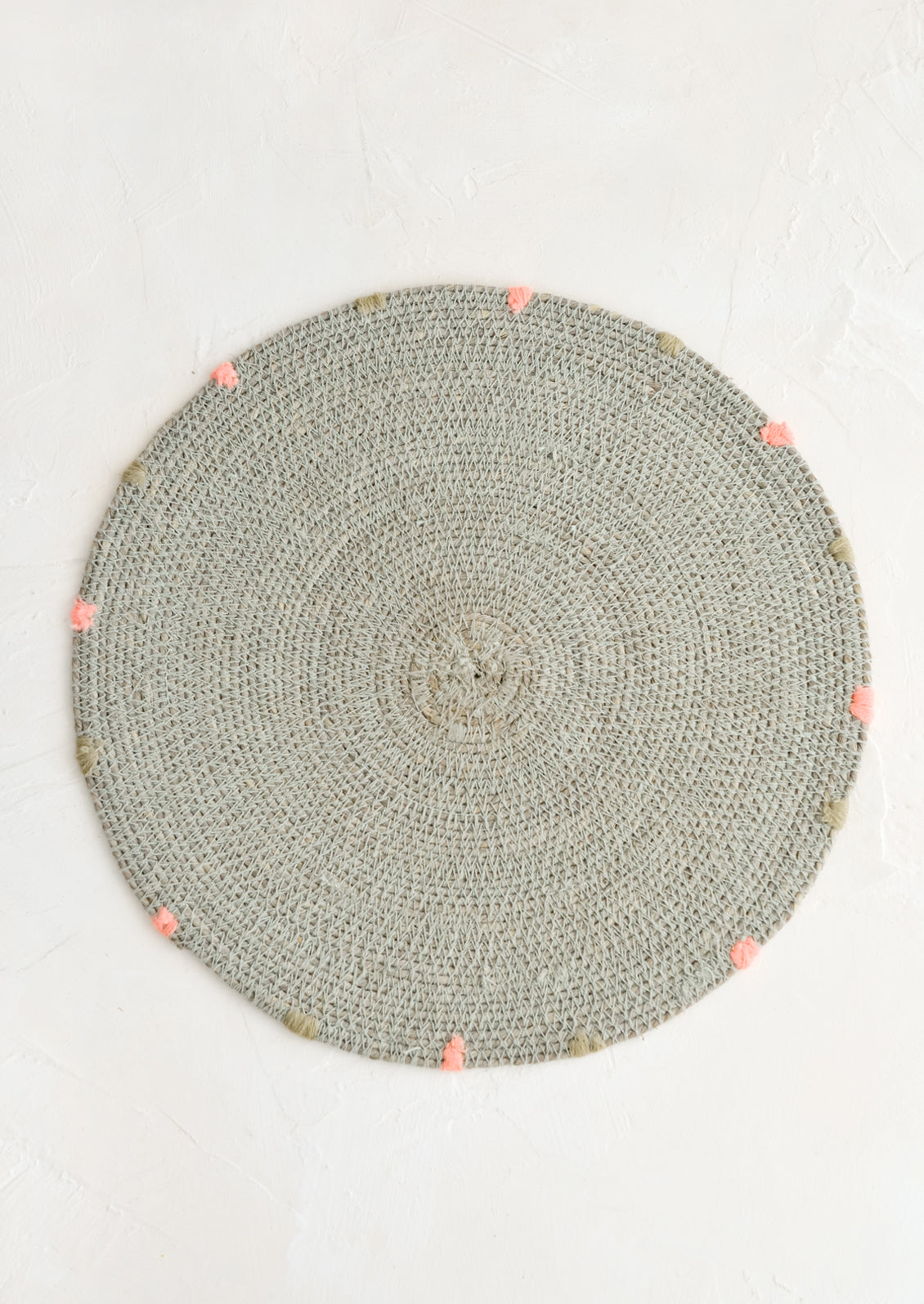 A round seagrass placemat in grey with grey and pink dashes embroidered around rim.