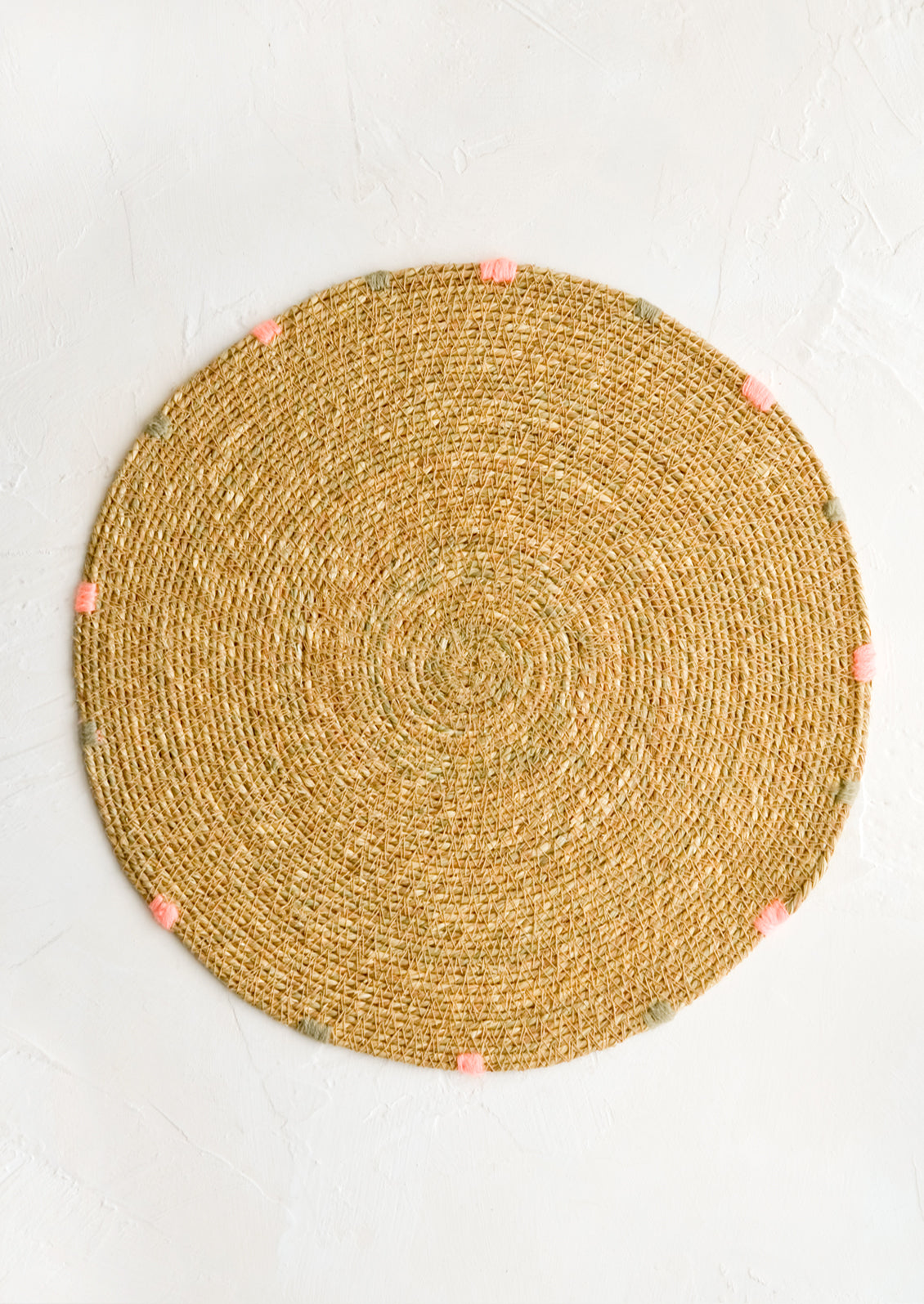 A round seagrass placemat in natural with grey and pink dashes embroidered around rim.