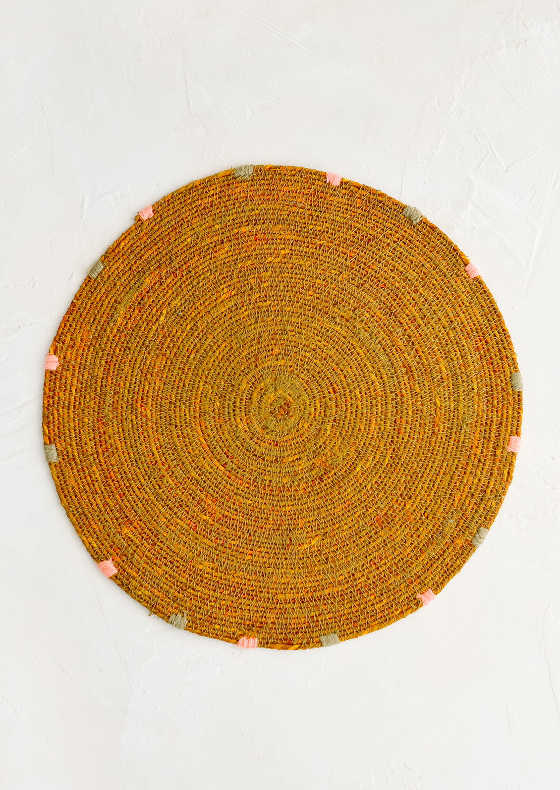 A round seagrass placemat in ochre with grey and pink dashes embroidered around rim.