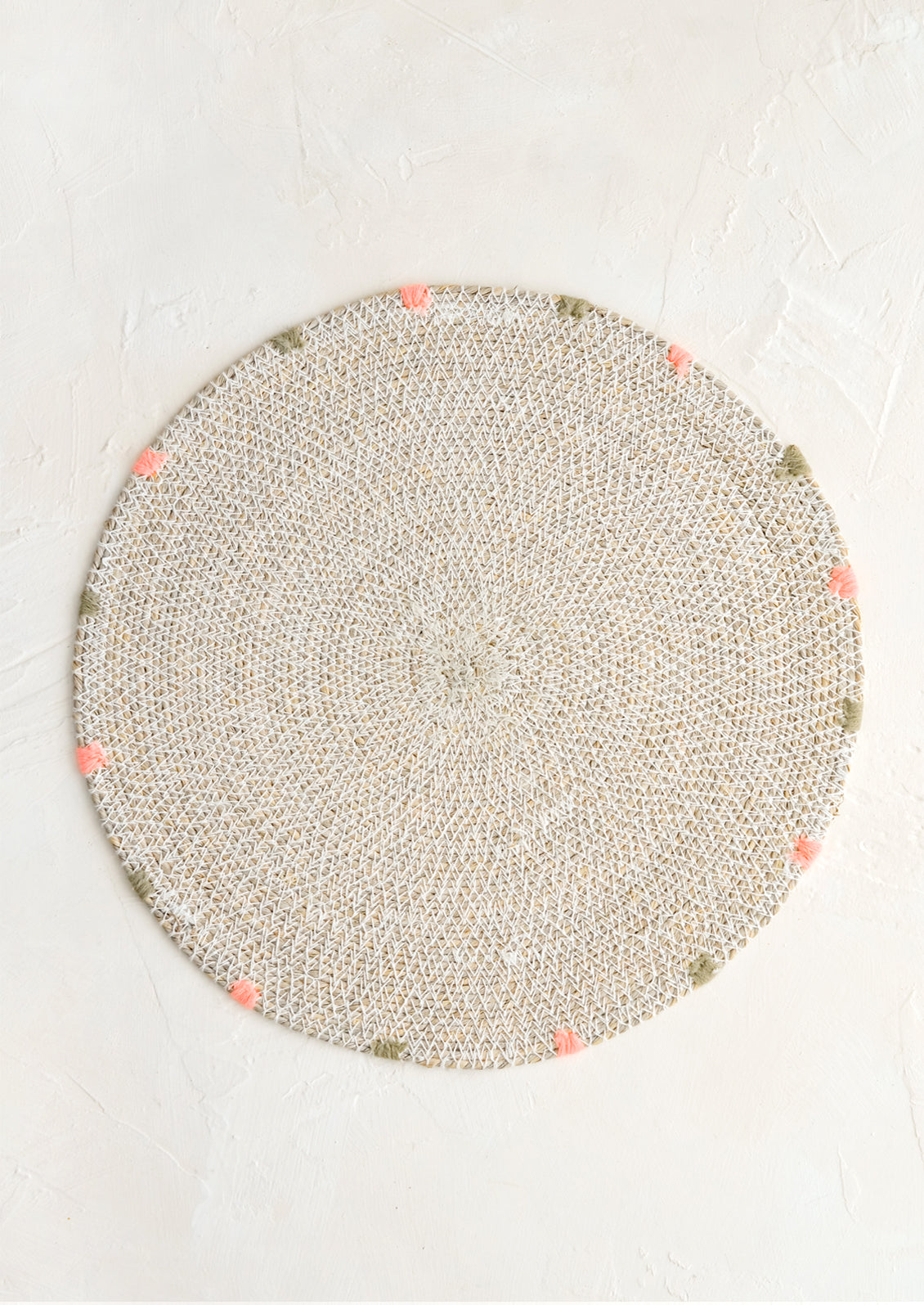 A round seagrass placemat in white with grey and pink dashes embroidered around rim.