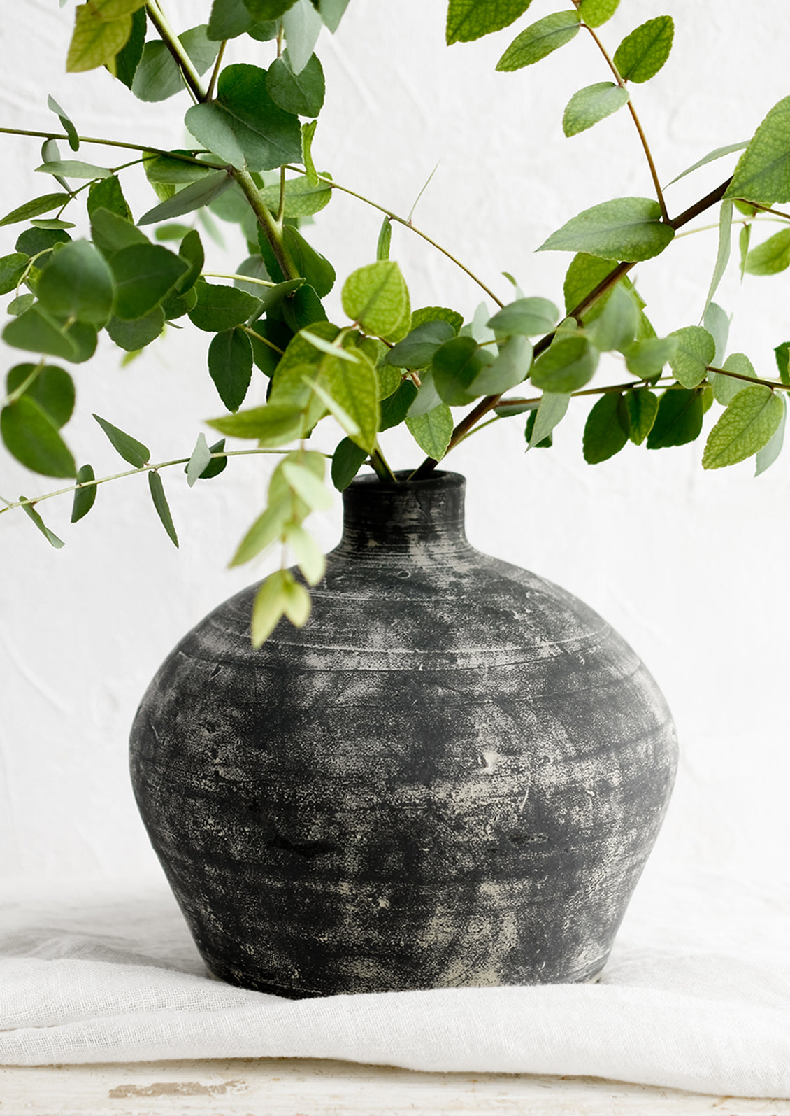A distressed black clay vase with narrow opening, with eucalyptus branches.