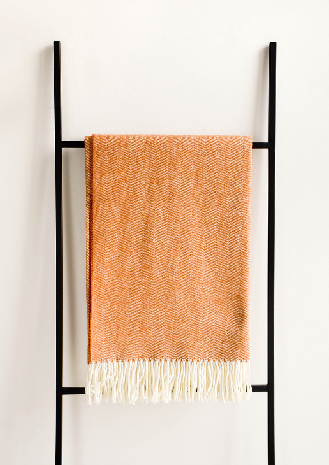 Soft blanket in colored herringbone weave. Ivory tassel trim at ends. Displayed hanging on a ladder.